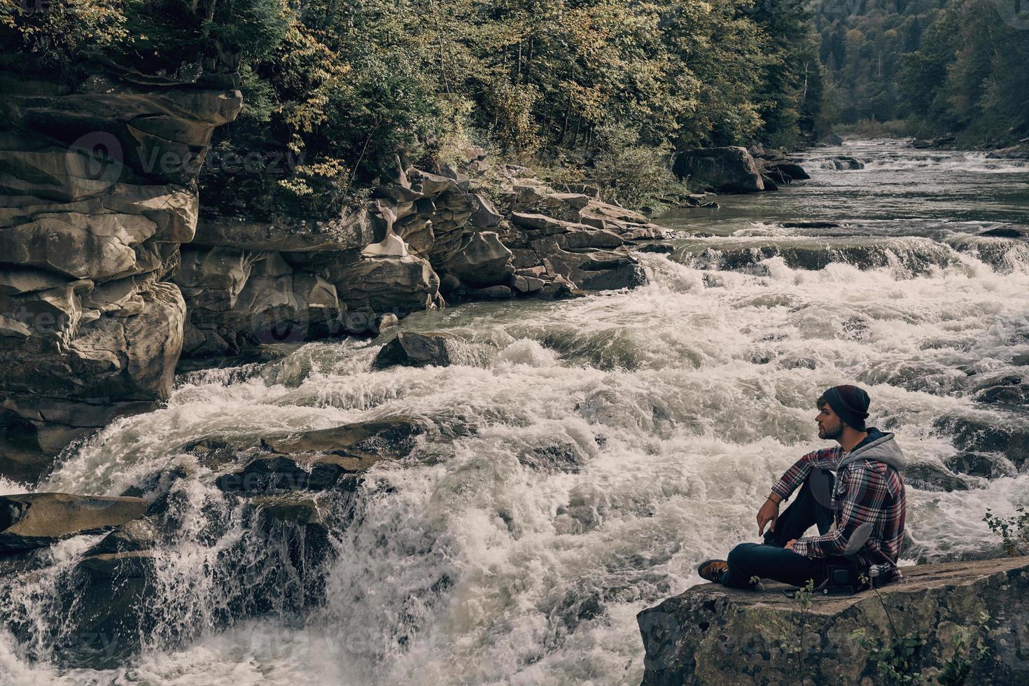 Ich würde für immer hier bleiben. hübscher junger moderner Mann, der wegschaut, während er auf dem Felsen in der Nähe des Flusses sitzt foto