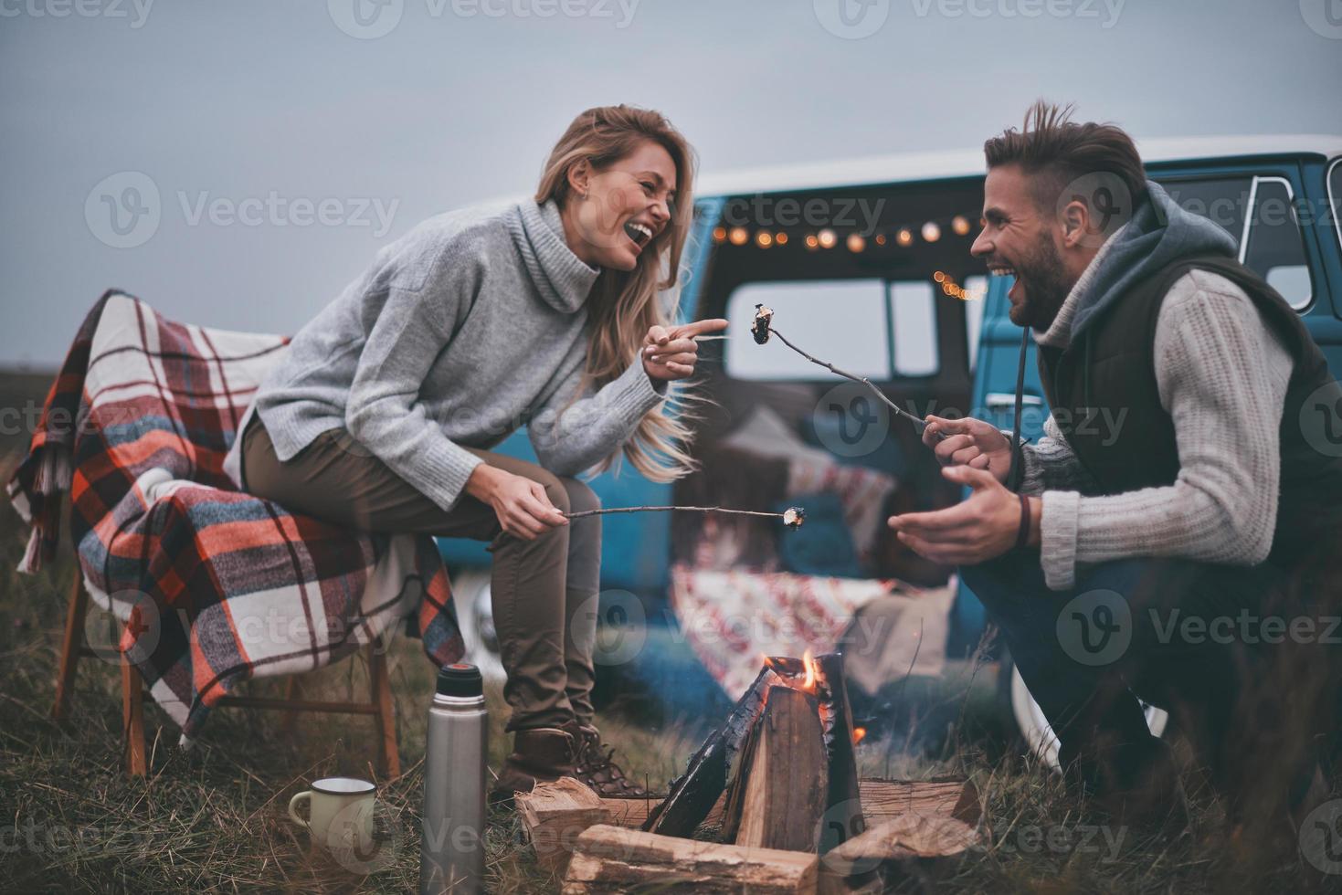Ausruhen nach einem langen Weg. Schönes junges Paar, das Marshmallows über einem Lagerfeuer röstet, während es seine Straßenreise genießt foto