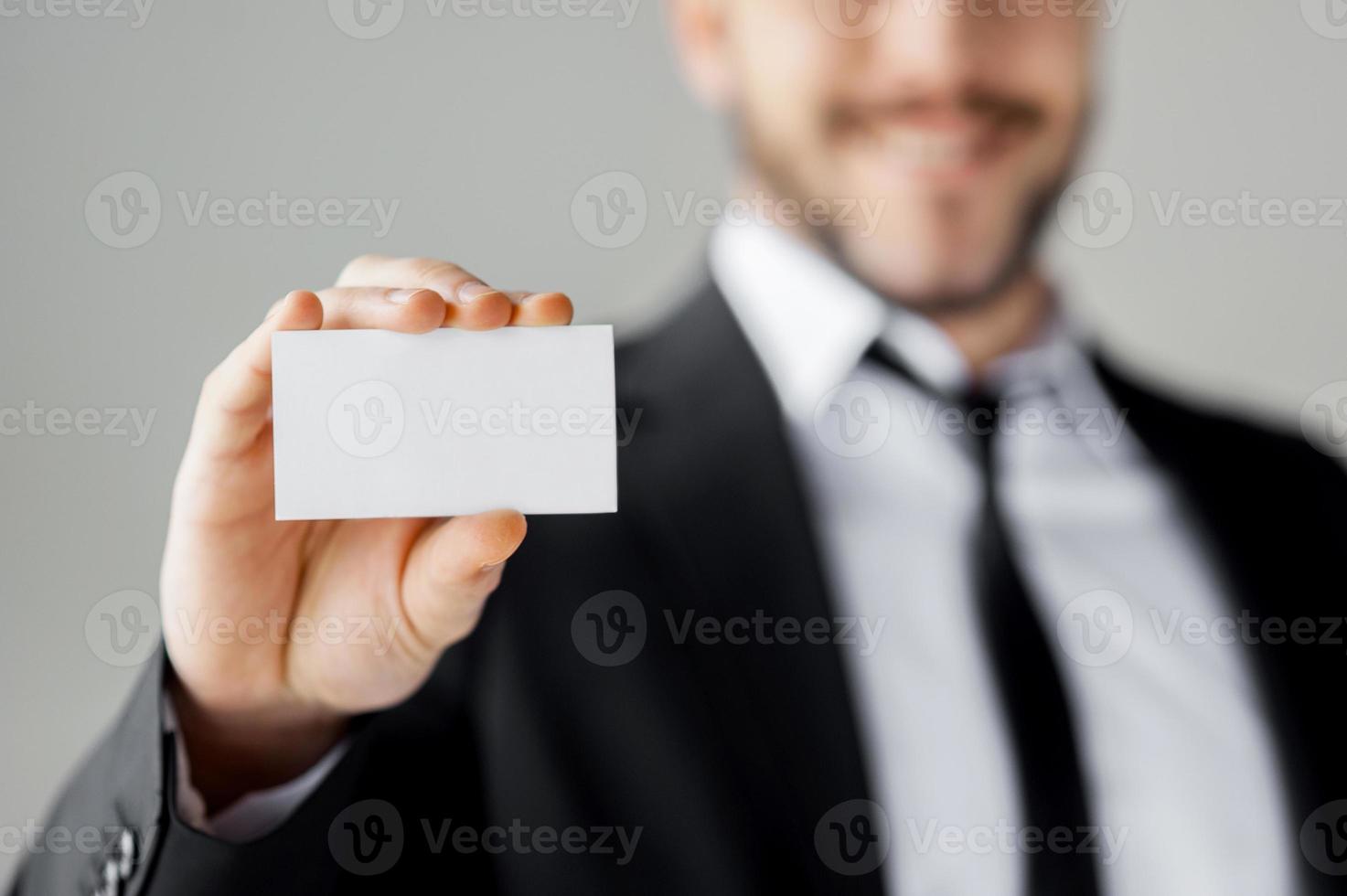 Kopieren Sie Platz auf seiner Visitenkarte. fröhlicher junger Mann in der Abendgarderobe, der seine Visitenkarte zeigt und lächelt, während er gegen grauen Hintergrund steht foto