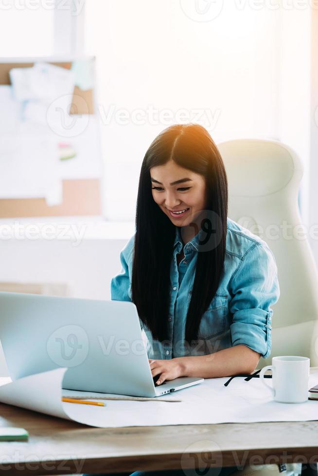 genießt ihren Arbeitstag. schöne junge asiatische frau, die laptop mit lächeln beim sitzen an ihrem arbeitsplatz verwendet foto