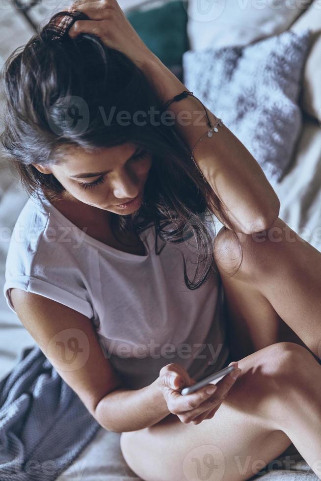 SMS an Freund. attraktive junge frau, die smartphone benutzt und hand im haar hält, während sie zu hause auf dem bett sitzt foto