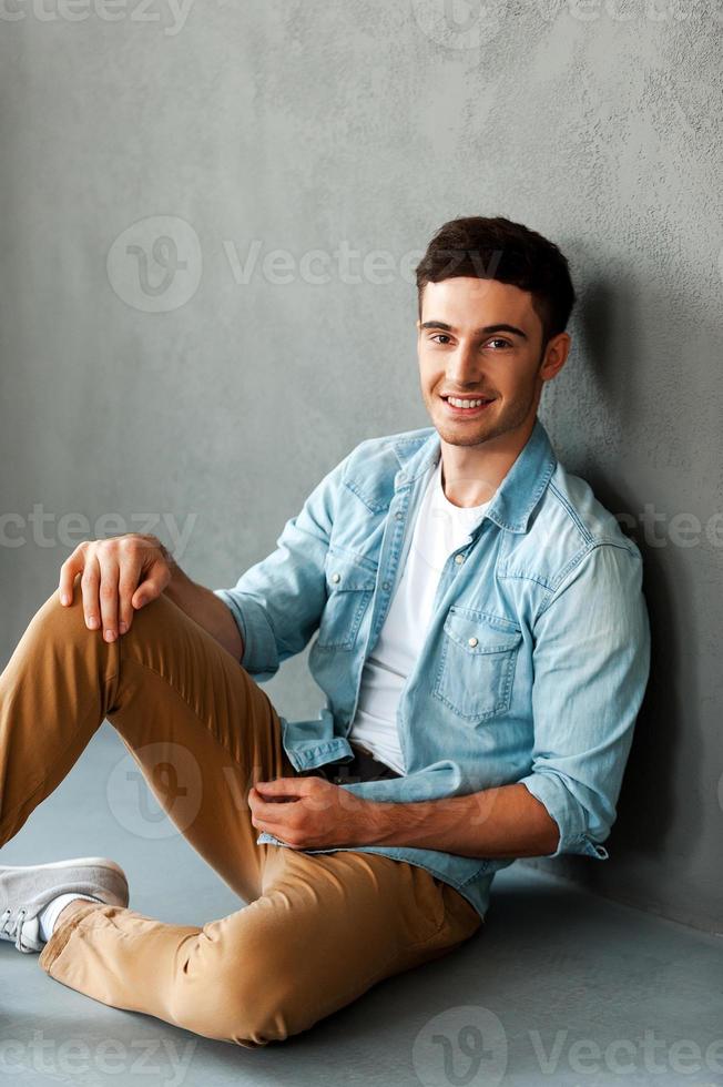 fröhlich und gutaussehend. glücklicher junger mann, der kamera betrachtet und beim sitzen gegen grauen hintergrund lächelt foto