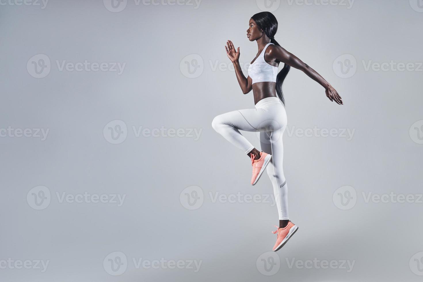 volle Länge der selbstbewussten jungen Afrikanerin in Sportkleidung, die vor grauem Hintergrund springt foto