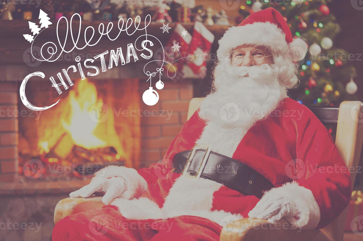 frohe weihnachten traditioneller weihnachtsmann, der an seinem stuhl sitzt und mit kamin und weihnachtsbaum im hintergrund lächelt foto