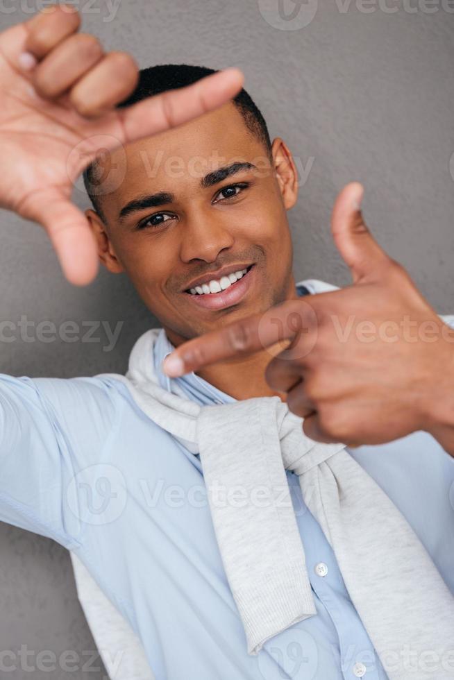 Konzentrieren Sie sich auf mich hübscher junger afrikanischer Mann, der einen Fingerrahmen macht und ihn mit einem Lächeln durchschaut, während er vor grauem Hintergrund steht foto