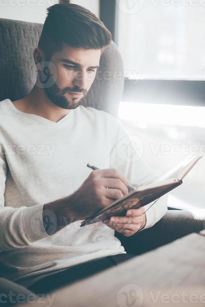 Notizen machen. Selbstbewusster junger Mann, der etwas in ein Notizbuch schreibt, während er an seinem Arbeitsplatz im Büro sitzt foto