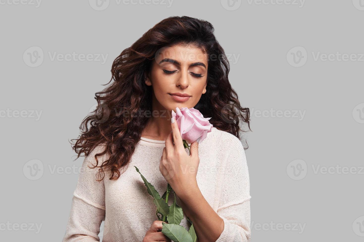 verloren in Träumen. Attraktive junge lächelnde Frau, die die Augen geschlossen hält und eine Blume hält, während sie vor grauem Hintergrund steht foto