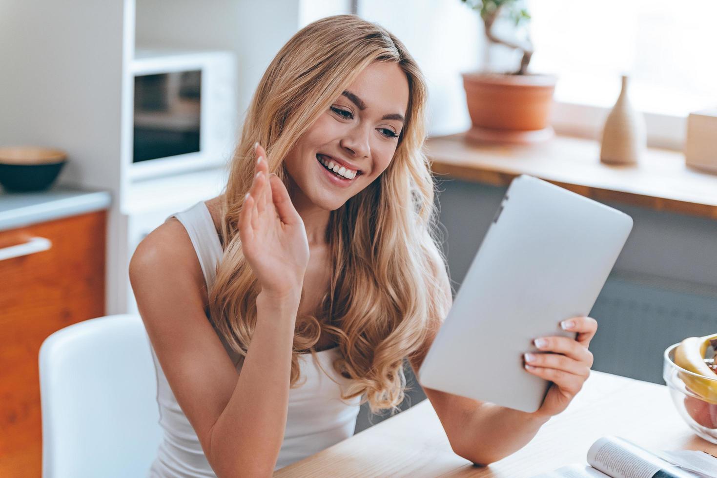 Entfernung ist kein Problem. Schöne, fröhliche junge Frau, die ihr Touchpad hält und lächelnd mit der Hand winkt, während sie zu Hause in der Küche sitzt foto