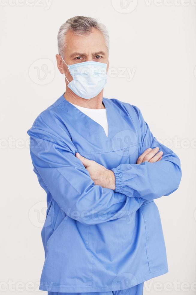 Sie können mir vertrauen. leitender Arzt mit grauem Haar in chirurgischer Maske, der in die Kamera schaut und die Arme verschränkt hält, während er isoliert auf Weiß steht foto