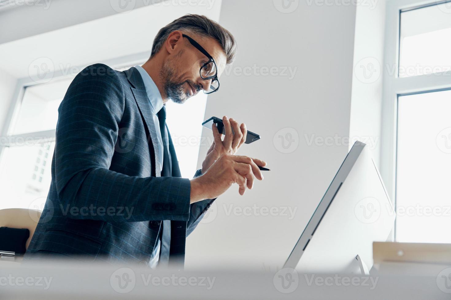 Schöner Geschäftsmann, der Lautsprecher am Telefon benutzt, während er im Büro auf einen Computermonitor zeigt foto