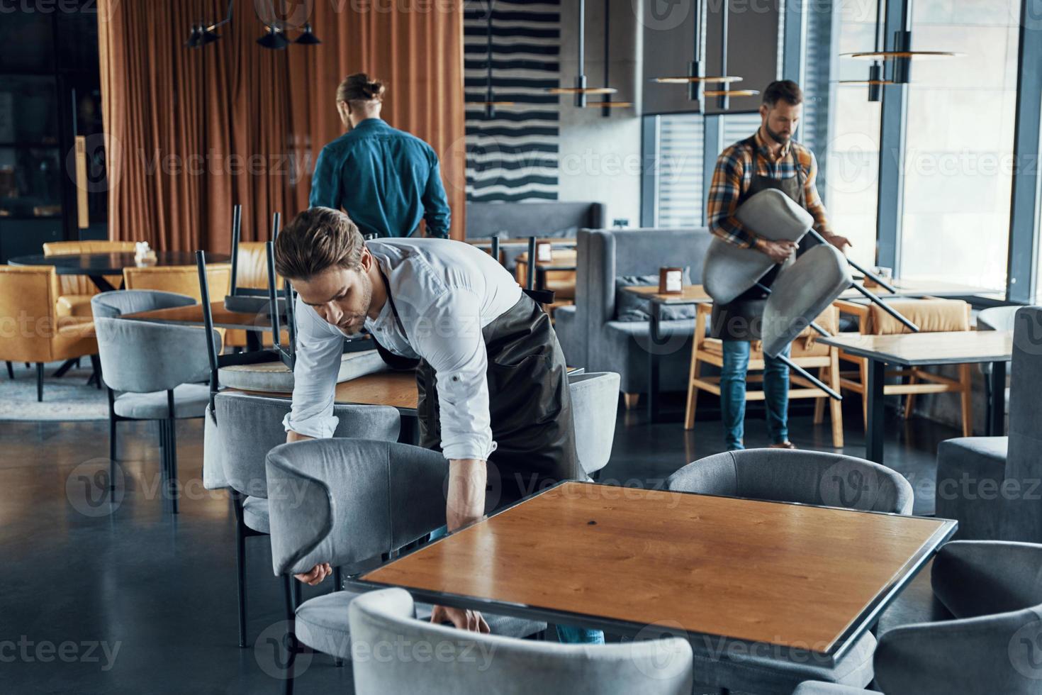 Hübsche junge Männer in Schürzen, die Möbel arrangieren, während sie das Restaurant auf die Eröffnung vorbereiten foto