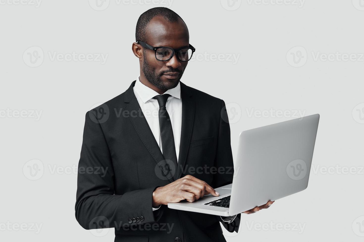 Beschäftigter junger afrikanischer Mann in Abendkleidung, der mit dem Computer arbeitet, während er vor grauem Hintergrund steht foto