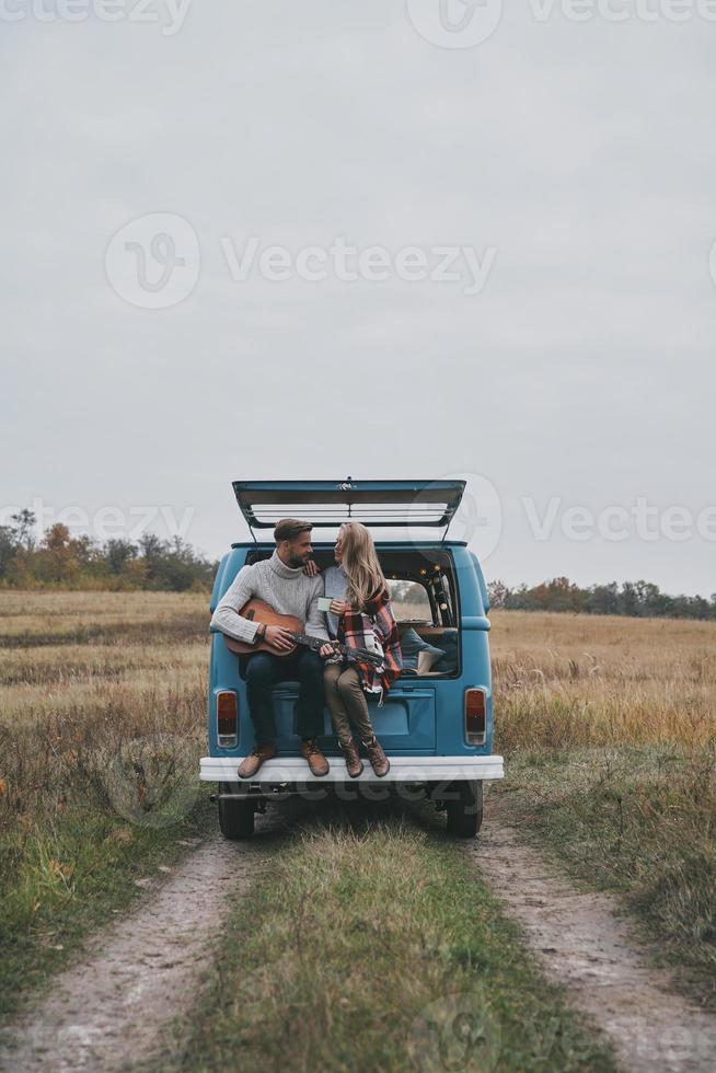 beste Reise. hübscher junger mann, der gitarre für seine schöne freundin spielt, während er im kofferraum des blauen minivans im retro-stil sitzt foto