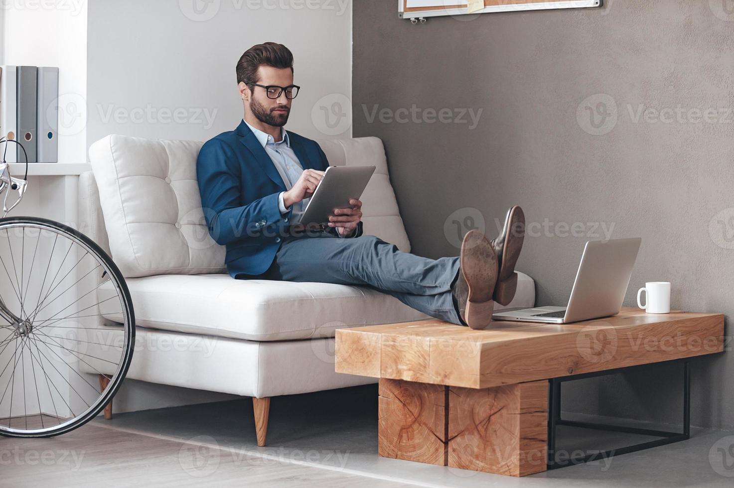 mit Freude arbeiten. hübscher junger Mann, der die Beine auf dem Tisch hält und mit dem Touchpad arbeitet, während er auf der Couch im Büro sitzt foto
