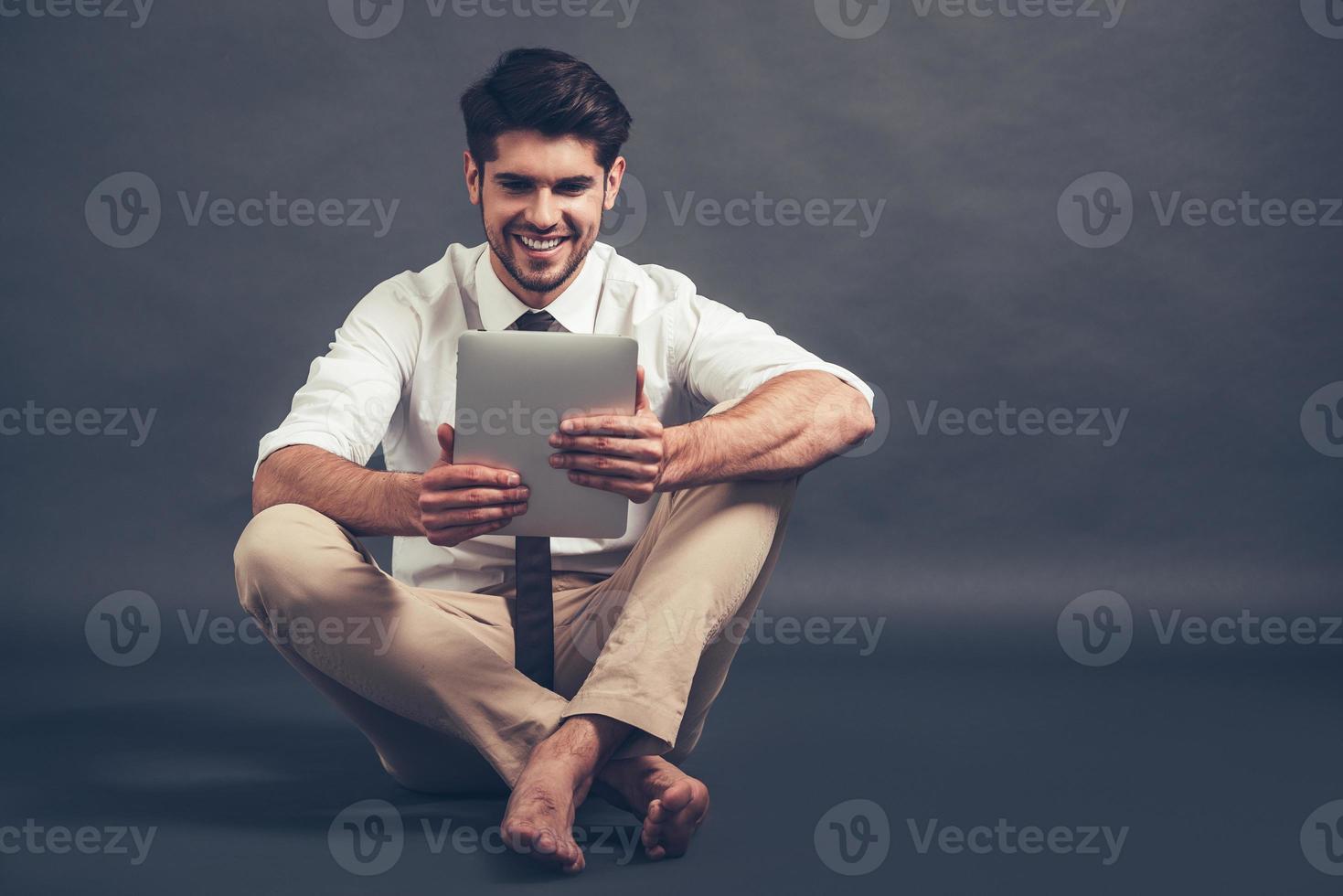 mit Freude arbeiten. voller Länge von selbstbewusstem jungen gutaussehenden Mann, der sein digitales Tablet mit einem Lächeln benutzt, während er auf dem Boden vor grauem Hintergrund sitzt foto