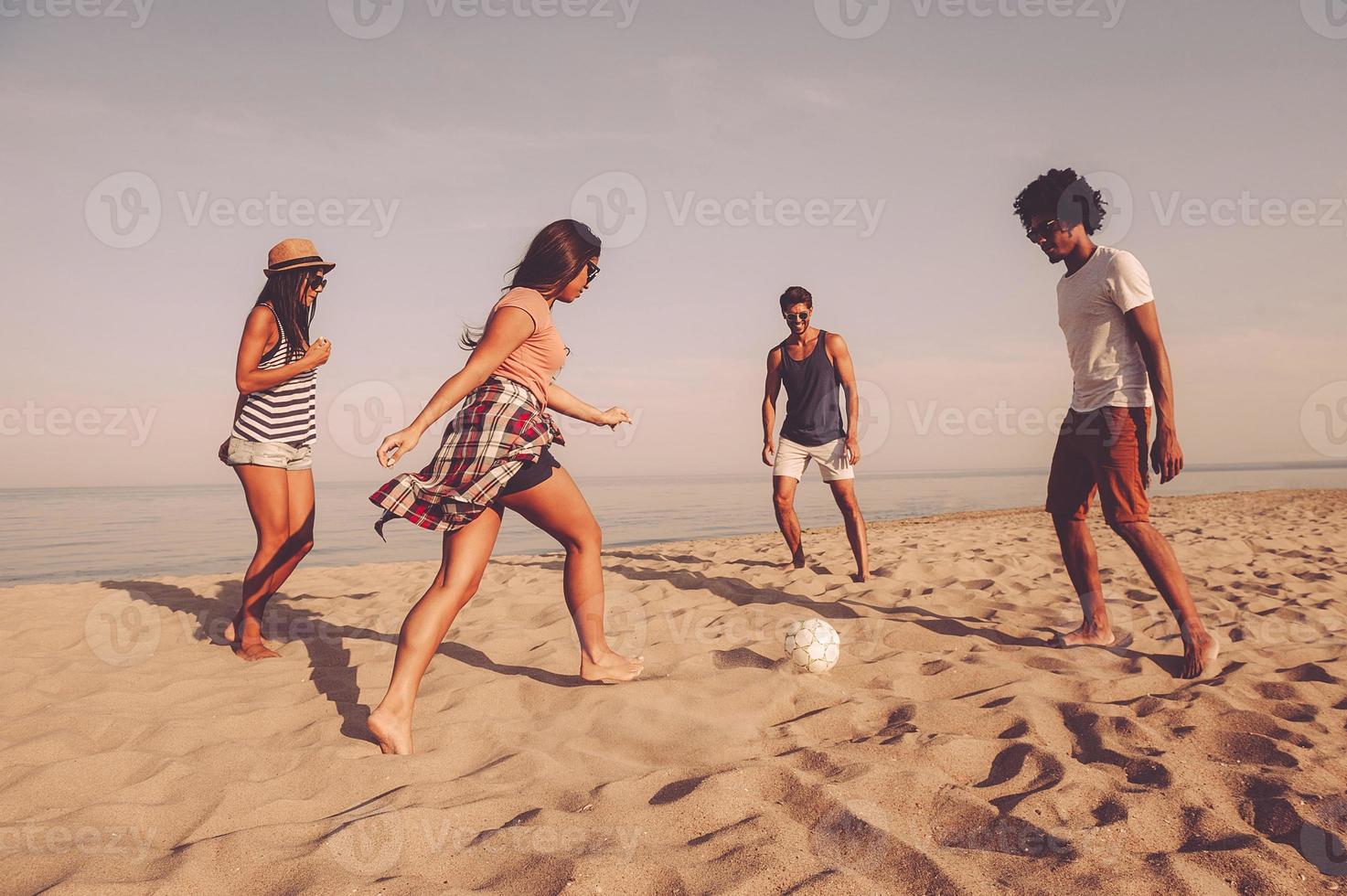 unbeschwerte gemeinsame Zeit verbringen. gruppe fröhlicher junger leute, die mit fußball am strand mit meer im hintergrund spielen foto