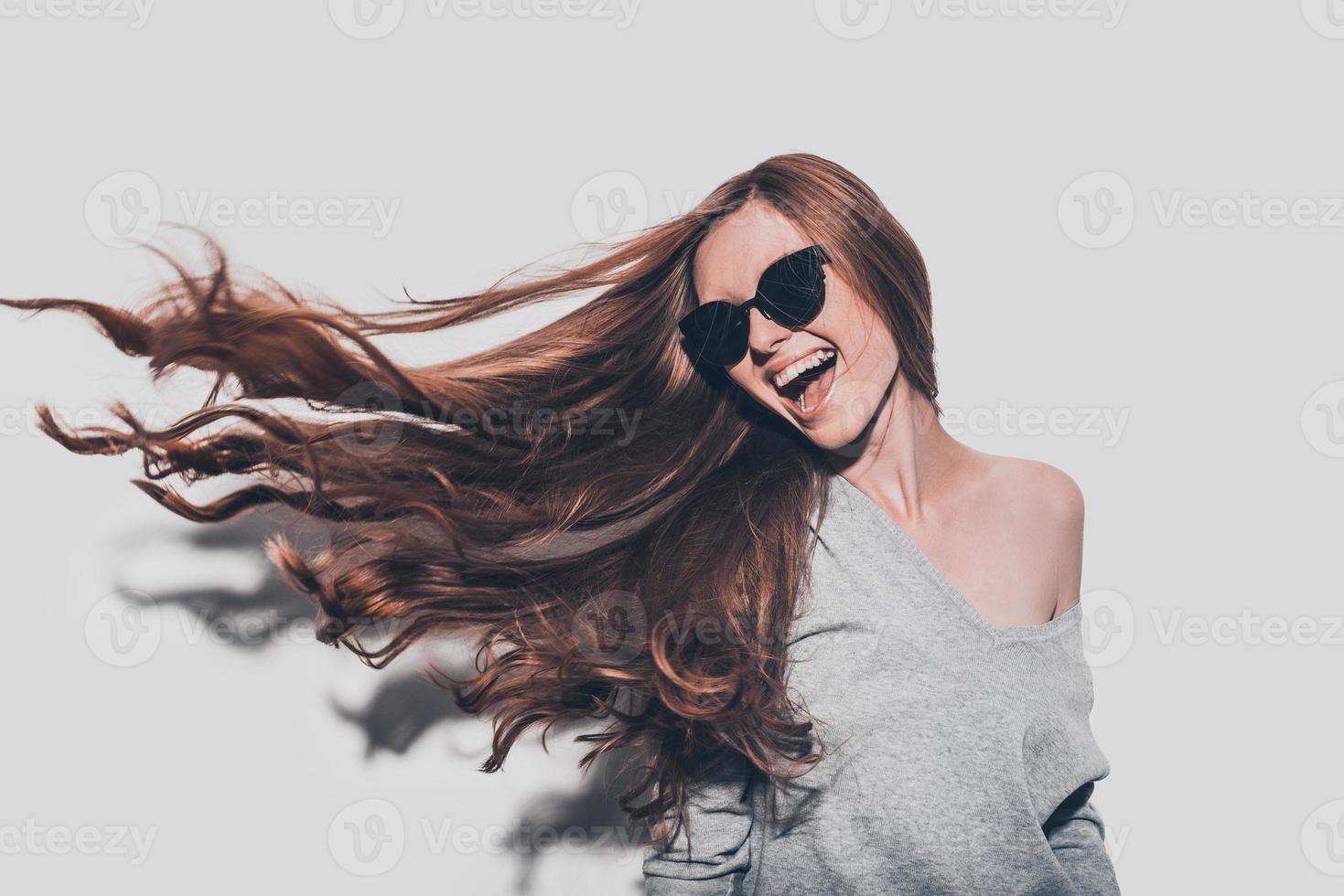 Haare wie Feuer. Attraktive junge lächelnde Frau mit Sonnenbrille und zerzaustem Haar, die wegschaut, während sie vor grauem Hintergrund steht foto