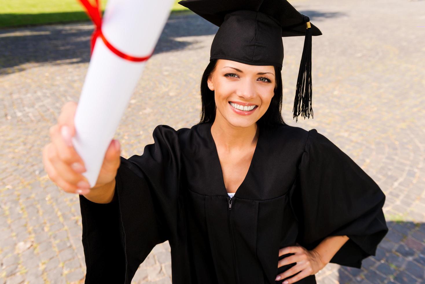 glücklicher Absolvent mit Diplom. Draufsicht einer glücklichen jungen Frau im Abschlusskleid, die ihr Diplom zeigt und lächelt foto