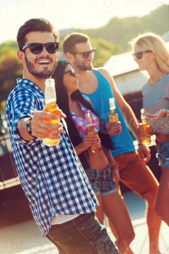 Begleiten Sie uns fröhlicher junger Mann, der eine Flasche Bier ausstreckt und in die Kamera schaut, während drei Personen im Hintergrund miteinander sprechen foto