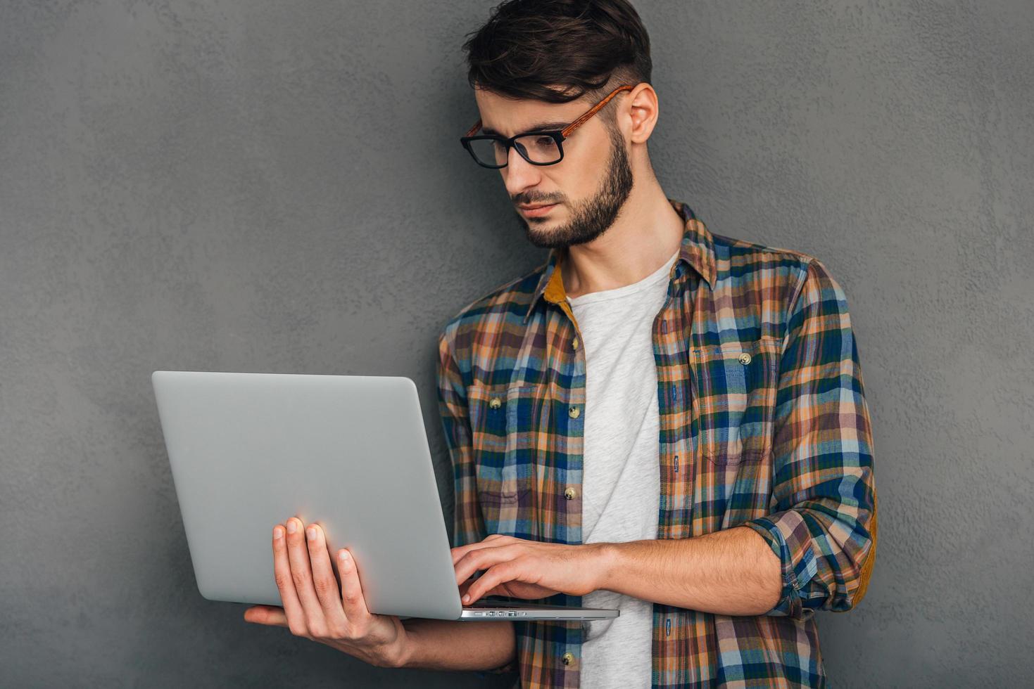 Überprüfung aller Updates. Selbstbewusster junger Mann, der seinen Laptop verwendet und konzentriert aussieht, während er vor grauem Hintergrund steht foto