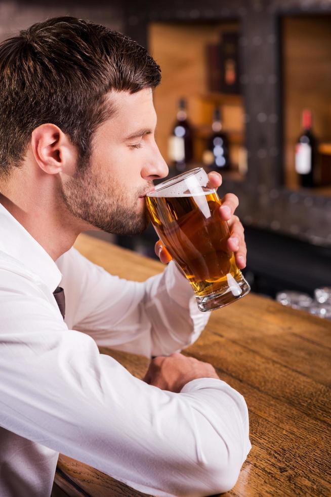 genießt sein Lieblingsbier. Seitenansicht eines hübschen jungen Mannes, der Bier trinkt, während er an der Bartheke sitzt foto