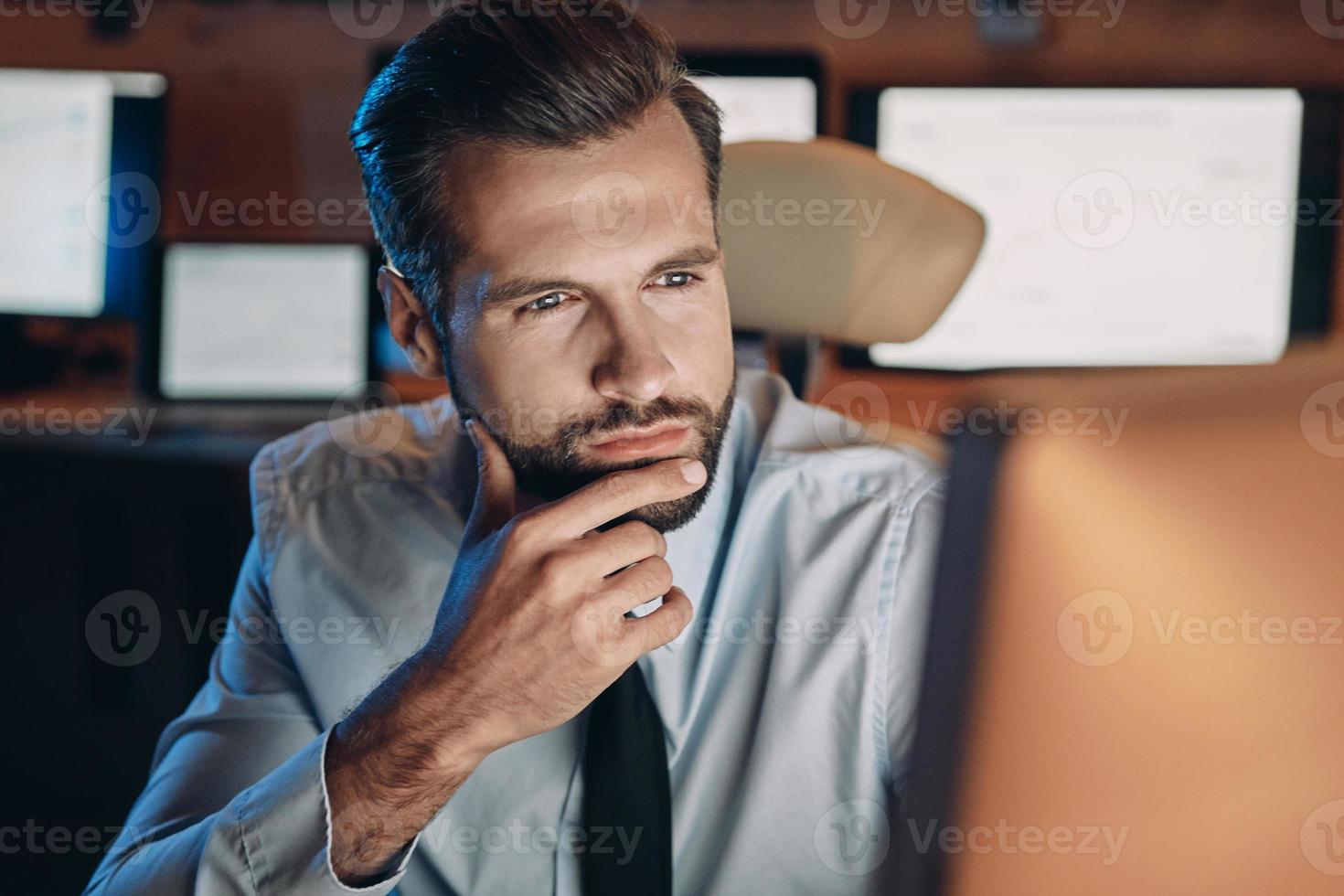 nachdenklicher junger Mann, der auf den Computer schaut, während er lange im Büro bleibt foto