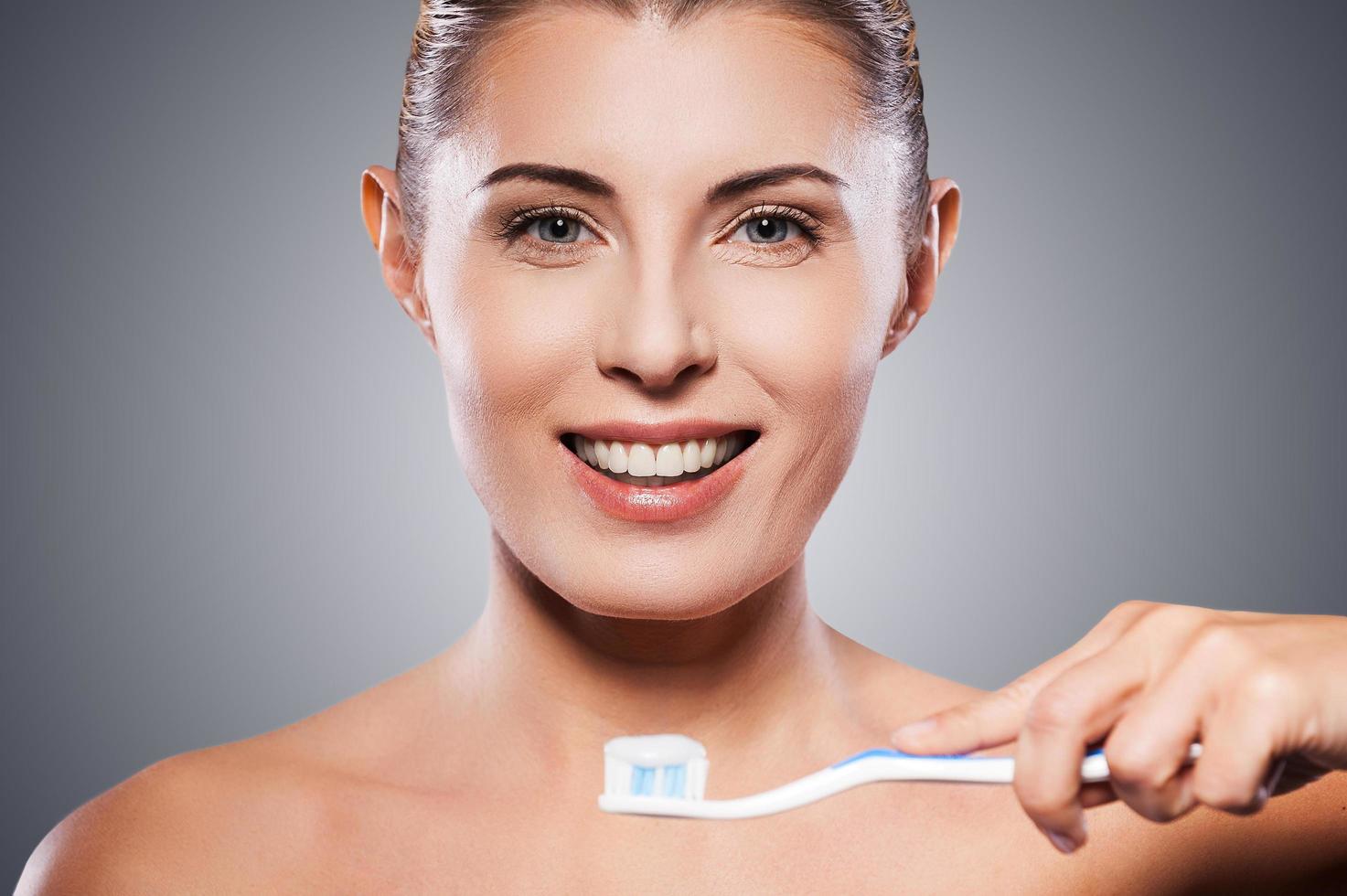 Zahnpflege. Schöne reife Frau mit Zahnbürste in der Nähe ihres Mundes und lächelnd in die Kamera, während sie vor grauem Hintergrund steht foto