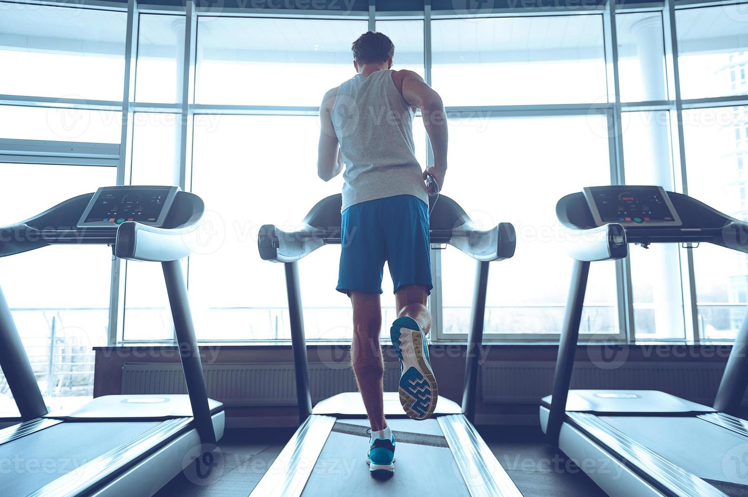 Joggen seinen Weg zu guter Gesundheit. Ganzkörperrückansicht eines jungen Mannes in Sportbekleidung, der auf dem Laufband vor dem Fenster im Fitnessstudio läuft foto