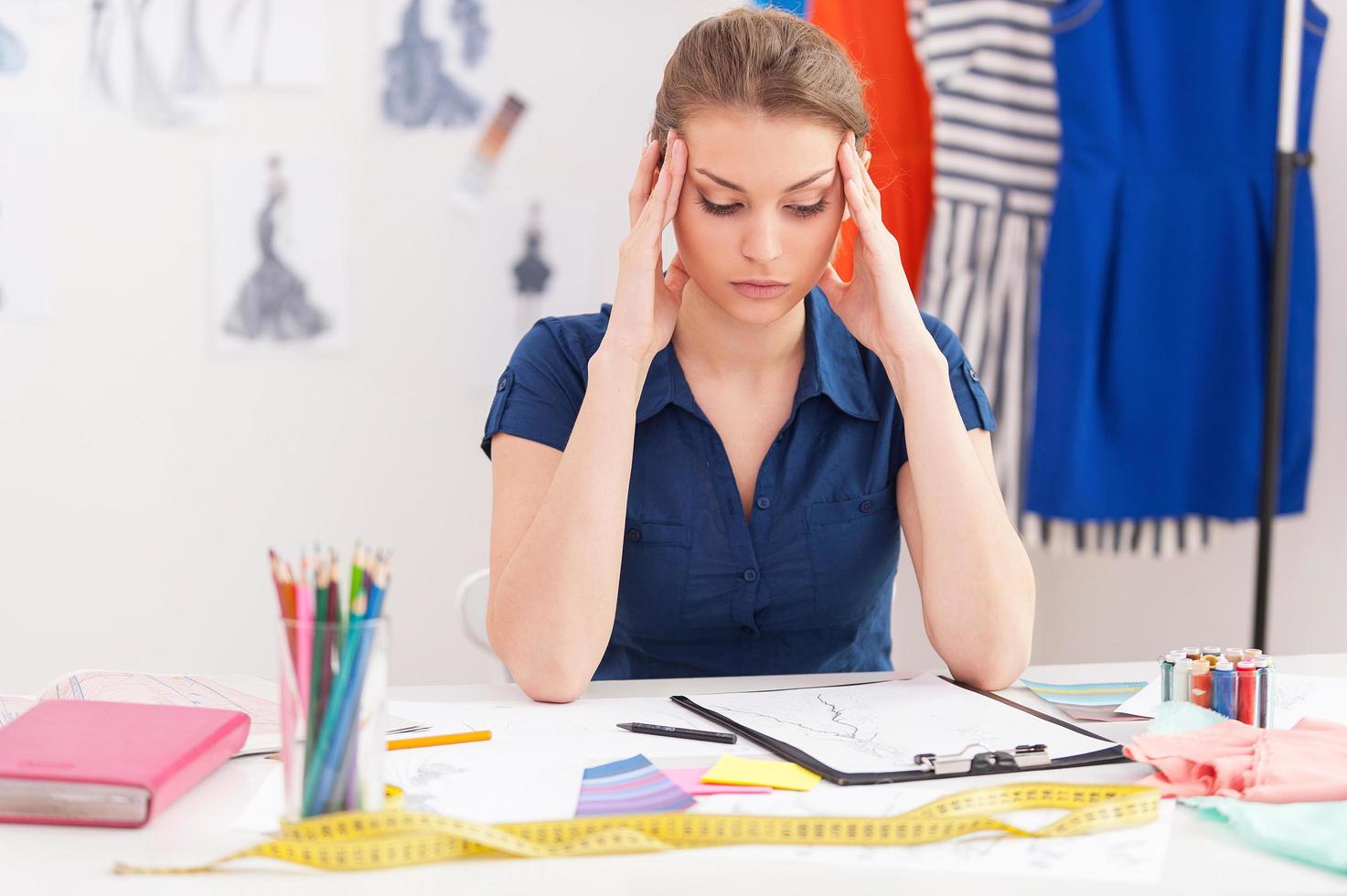 versuchen, die Frist zu erreichen. Depressive weibliche Modedesignerin, die den Kopf in den Händen hält, während sie an ihrem Arbeitsplatz sitzt foto