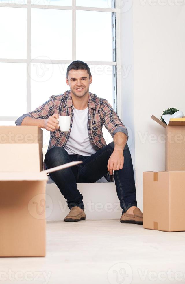 gerade in ein neues Haus gezogen. Fröhlicher junger Mann, der auf der Fensterbank sitzt und eine Kaffeetasse hält, während Kartons neben ihm liegen foto