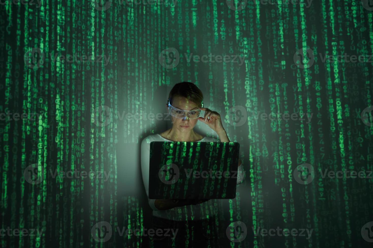 Selbstbewusste Frau, die eine futuristische Brille anpasst, während sie einen Laptop vor dunklem Hintergrund benutzt foto