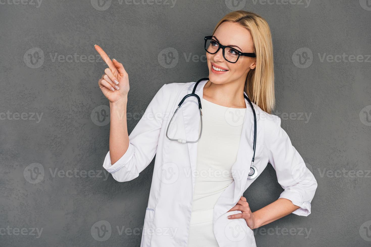 Der Hausarzt empfiehlt Ihnen eine schöne junge Ärztin, die auf den Kopierbereich zeigt und mit einem Lächeln wegschaut, während sie vor grauem Hintergrund steht foto