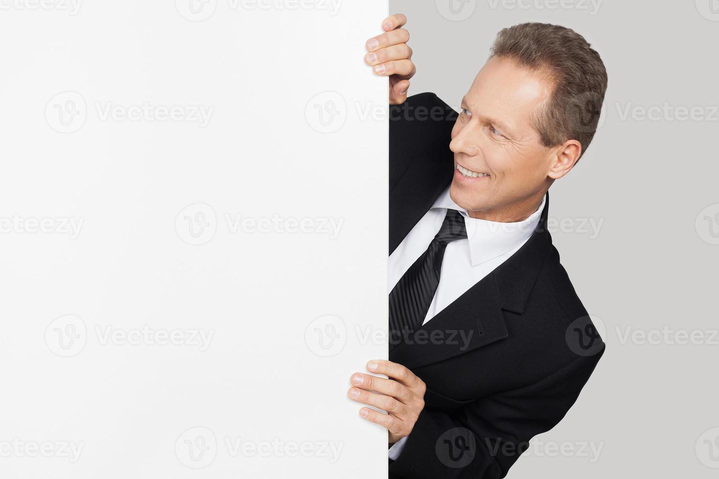 Blick auf deinen Text. Fröhlicher reifer Mann in Abendkleidung, der aus dem Kopierraum herausschaut und ihn betrachtet, während er vor grauem Hintergrund steht foto