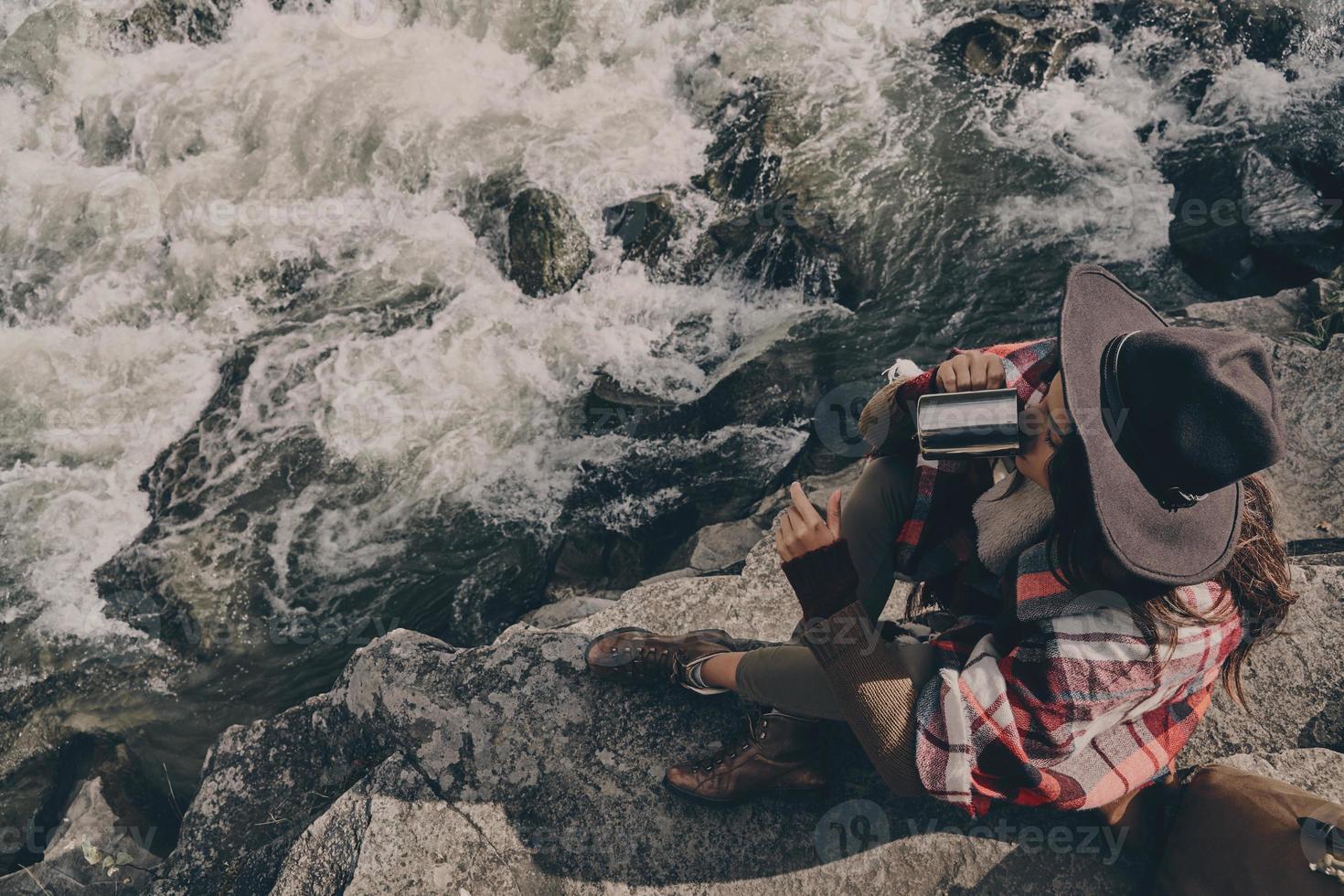 sich bereit machen, die Reise fortzusetzen. Blick von oben auf eine schöne junge Frau, die mit einer Decke bedeckt ist und heißes Tee trinkt, während sie in der Nähe des Flusses in den Bergen sitzt foto