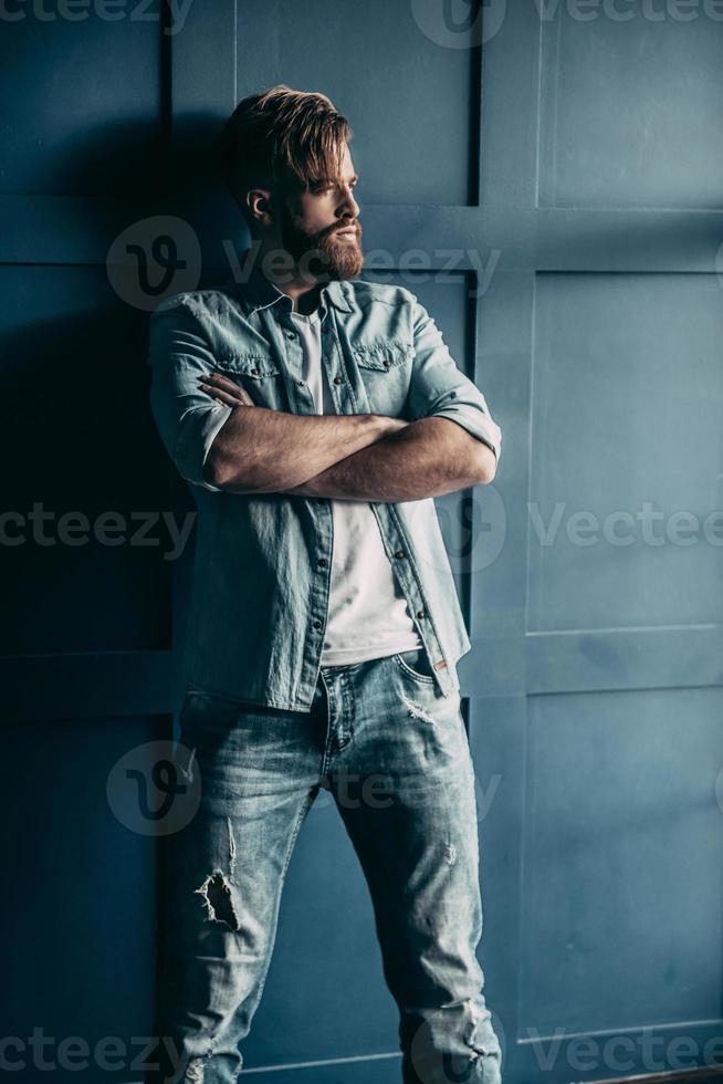 unwiderstehlicher Mann. Selbstbewusster junger bärtiger Mann im blauen Jeanshemd, der wegschaut und die Arme verschränkt hält, während er an der Wand steht foto