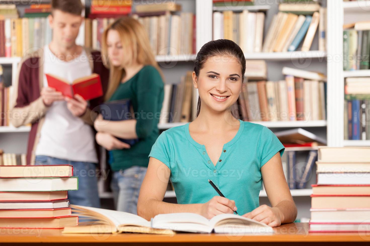 Ich werde zu meiner Abschlussprüfung bereit sein. Selbstbewusste junge Frau, die am Bibliothekstisch sitzt und lächelt, während zwei Personen im Hintergrund ein Buch lesen foto