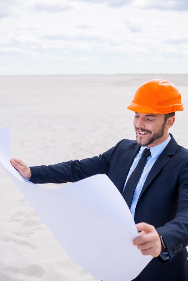 inspirierter Architekt. Fröhlicher junger Mann in Abendkleidung und Hardhat, der Blaupausen untersucht, während er in der Wüste steht foto