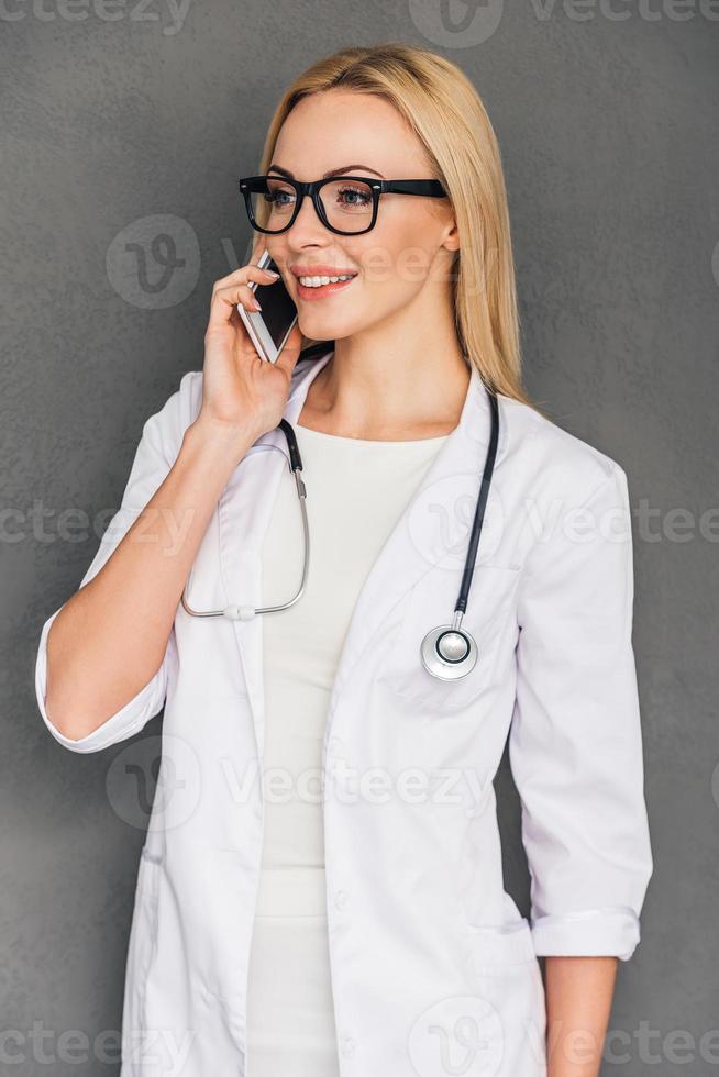 gutes Gespräch mit ihrer Patientin schöne junge Ärztin, die mit einem Lächeln am Handy spricht, während sie vor grauem Hintergrund steht foto