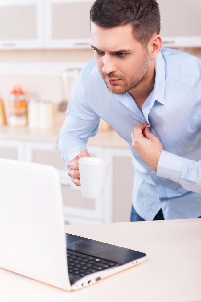 Morgenroutine. besorgter junger mann, der sein hemd anpasst und auf den laptop schaut, während er in der küche steht foto