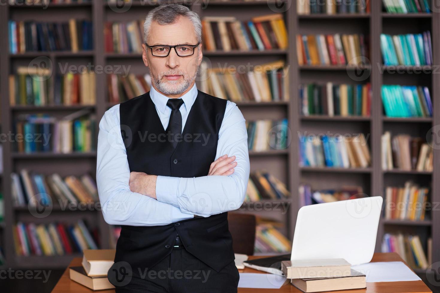 selbstbewusster Professor. Selbstbewusster älterer Mann mit grauem Haar in Abendkleidung, der die Arme verschränkt hält und in die Kamera schaut, während er sich am Tisch lehnt und mit einem Bücherregal im Hintergrund foto