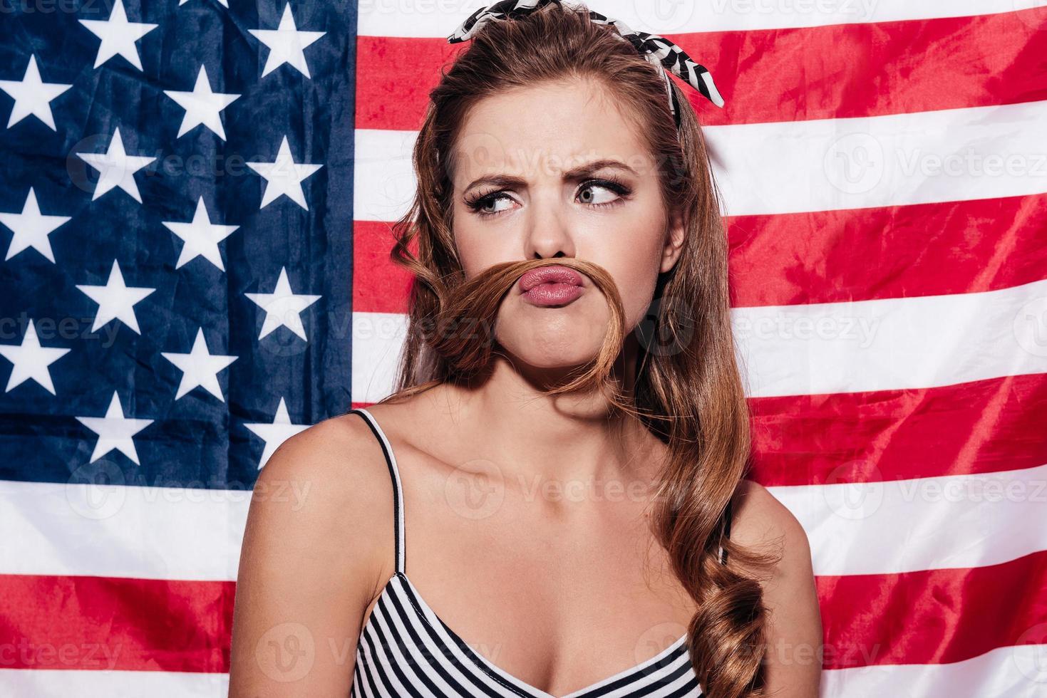 Schauen Sie sich mein Schnurrbartporträt einer jungen Frau an, die einen falschen Schnurrbart aussehen lässt, während sie gegen die amerikanische Nationalflagge steht foto