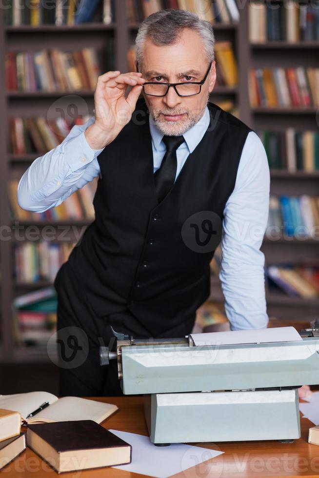 klug und selbstbewusst. Selbstbewusster älterer Mann mit grauem Haar in Abendkleidung, der sich mit Schreibmaschine darauf und Bücherregal im Hintergrund an den Tisch lehnt foto