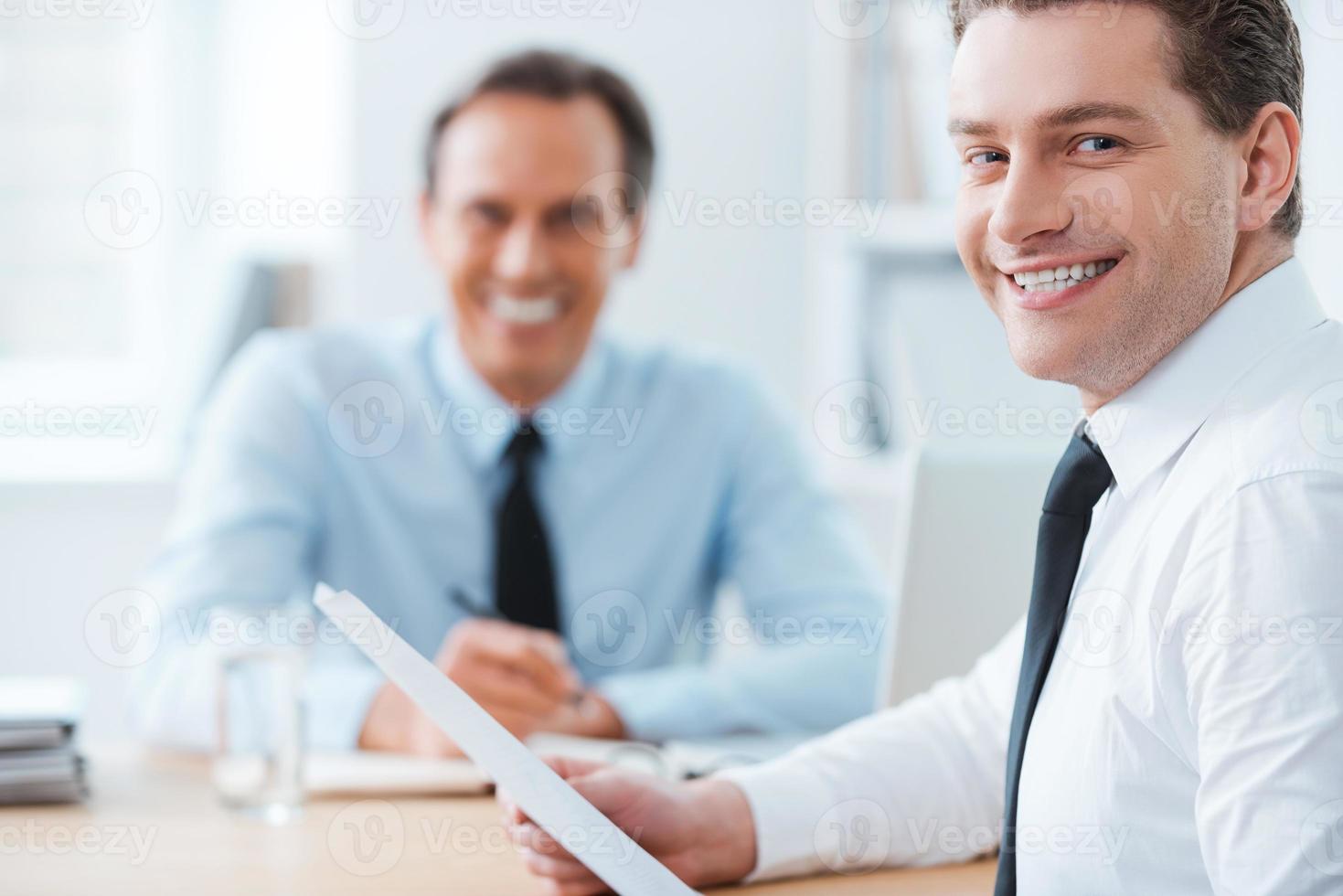 produktiver tag im büro. glücklicher junger geschäftsmann in formalwear, der über die schulter schaut, während sein chef im hintergrund sitzt foto
