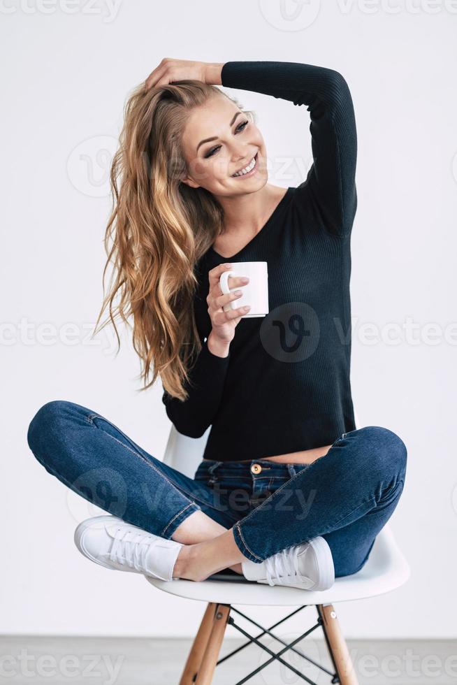 fröhliche Schönheit. schöne junge Frau, die Kaffeetasse hält und mit einem Lächeln wegschaut, während sie auf einem Stuhl im Lotussitz vor weißem Hintergrund sitzt foto