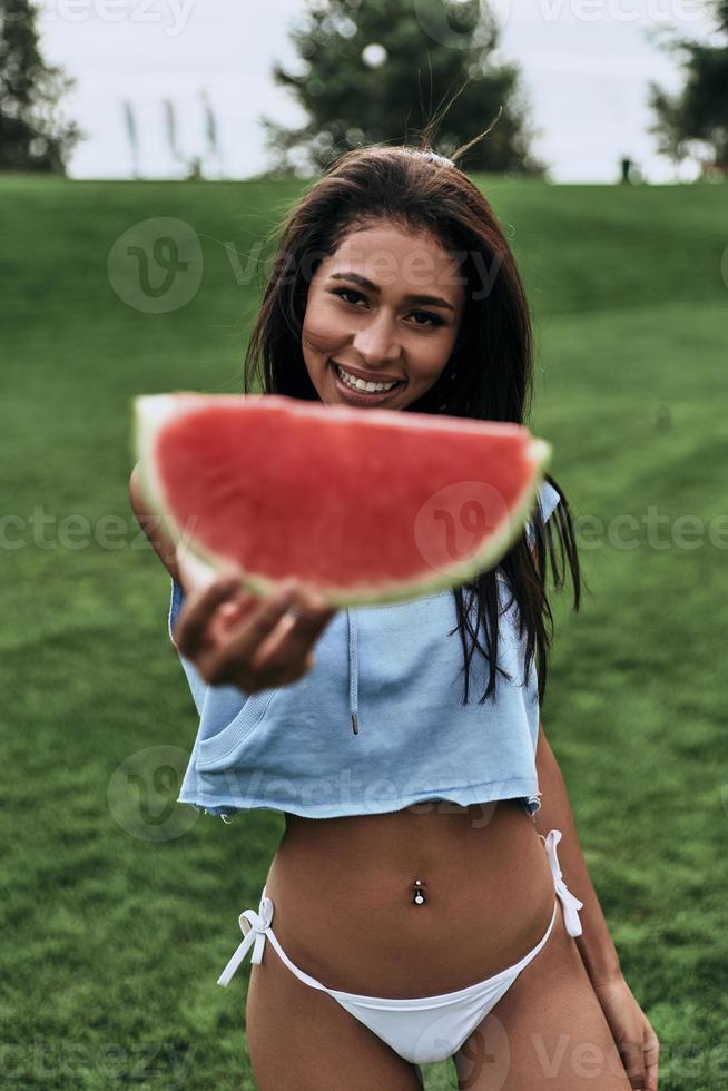 Attraktive junge Frau, die die Hand mit einer Scheibe Wassermelone ausstreckt und lächelt, während sie im Freien steht foto