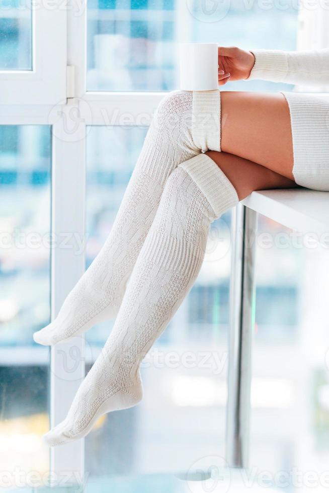 heißes Getränk zu Hause genießen. Nahaufnahme einer Frau in weißen warmen Socken, die eine Kaffeetasse hält, während sie zu Hause vor dem Fenster sitzt foto