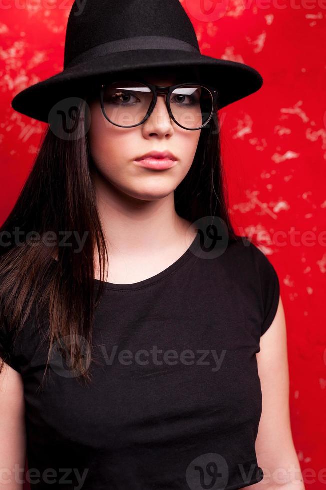 Schönheit im Stil. Studioaufnahme einer schönen jungen Frau mit Hut und Brille, die vor rotem Hintergrund steht foto