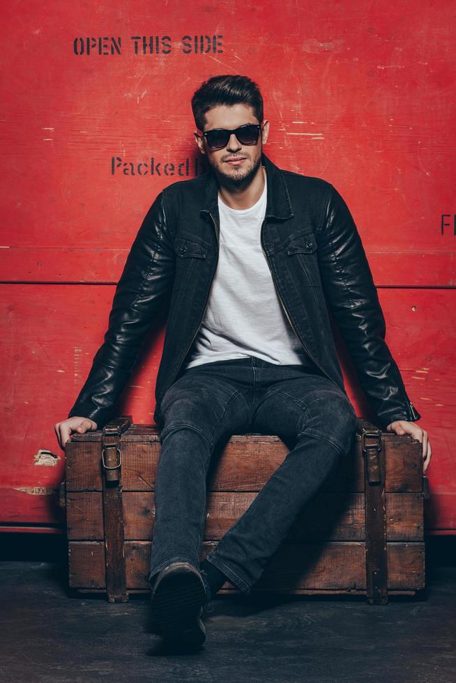 modern und ansehnlich. hübscher junger Mann, der seine Sonnenbrille anpasst, während er auf einer Holzkiste vor rotem Hintergrund sitzt foto