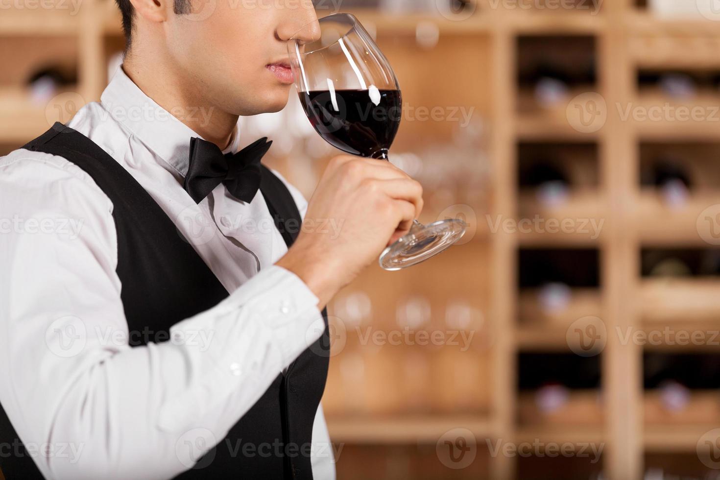 einen guten Wein riechen. Zugeschnittenes Bild eines selbstbewussten jungen Sommeliers, der mit Weinflaschen vor einem Regal steht und die Arme verschränkt hält foto