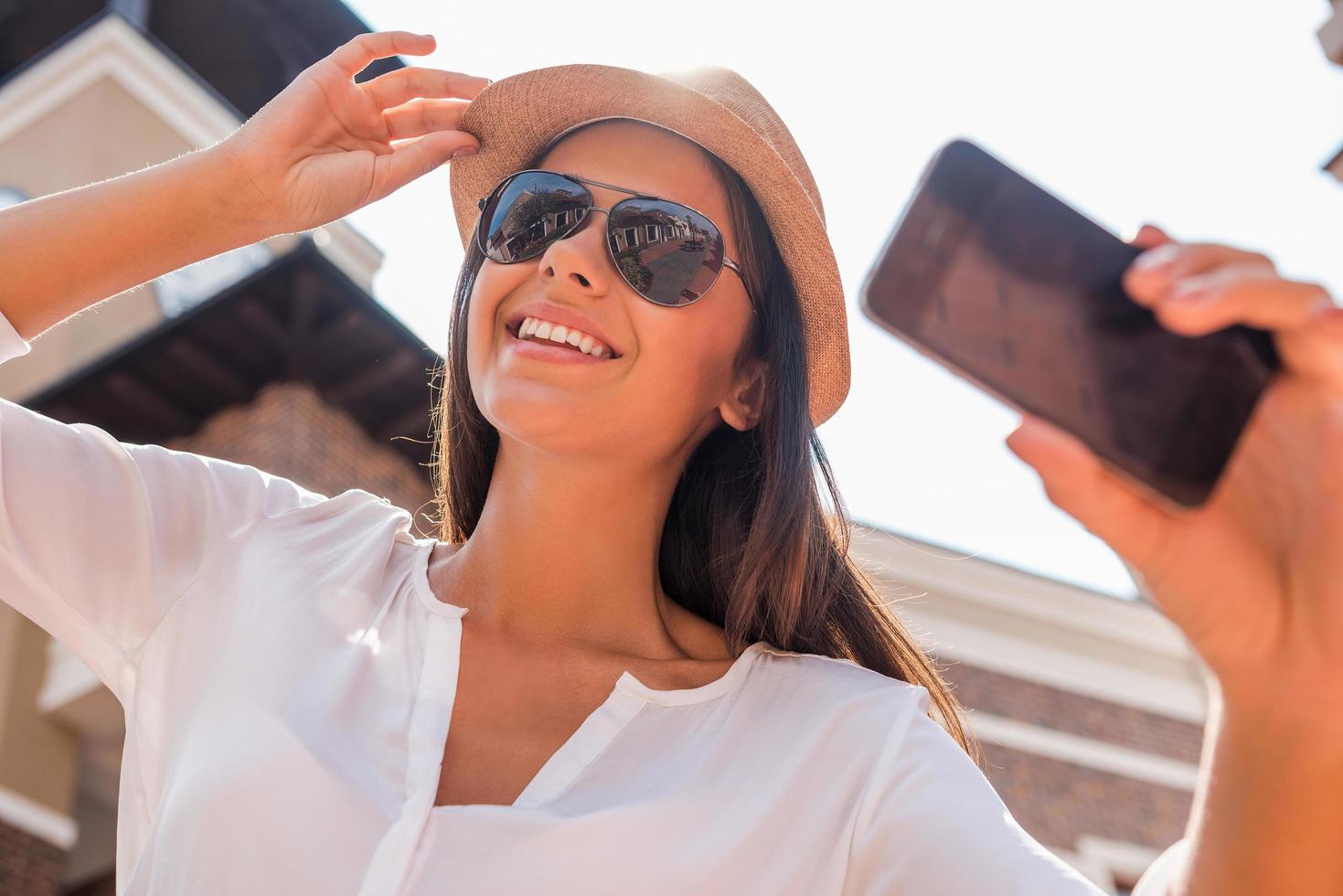 Selfie für meinen Freund. Tiefwinkelansicht einer schönen jungen flippigen Frau, die ihren Hut anpasst und ein Selfie macht, während sie im Freien steht foto